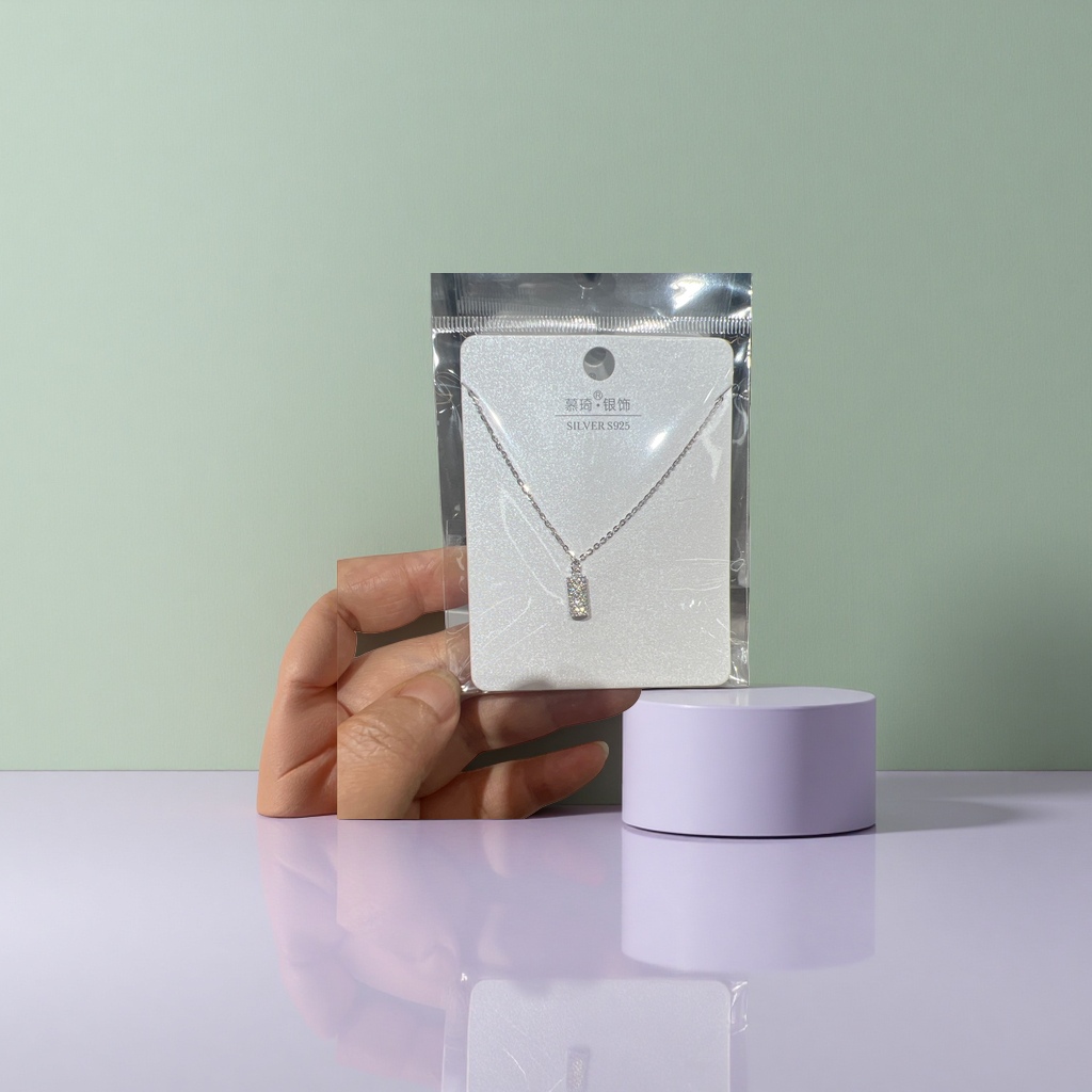 A silver full diamond rectangular pendant necklace with a fine chain, supported by white glitter cardstock for display, and packaged in transparent plastic details Picture