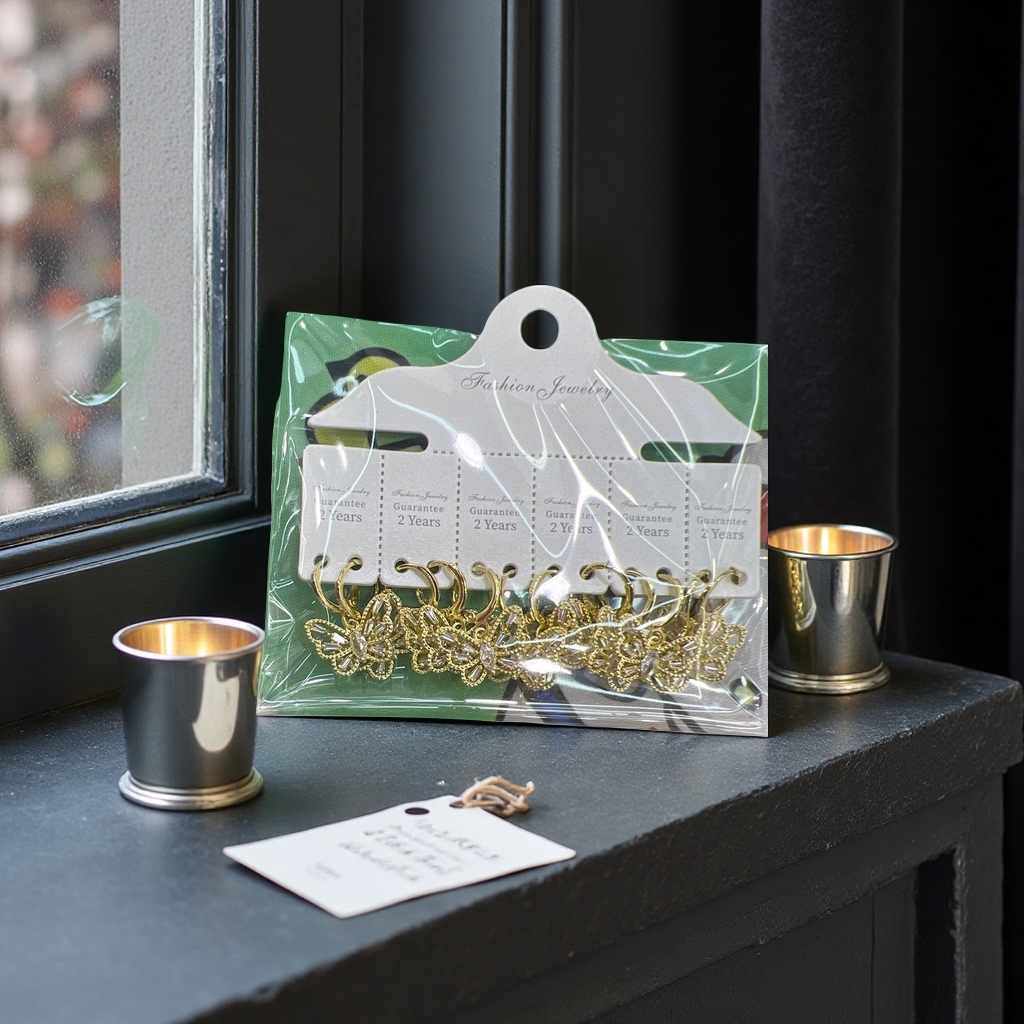 Golden butterfly-shaped earrings, packaged in a transparent plastic bag, with a white tag and a green cartoon fabric background