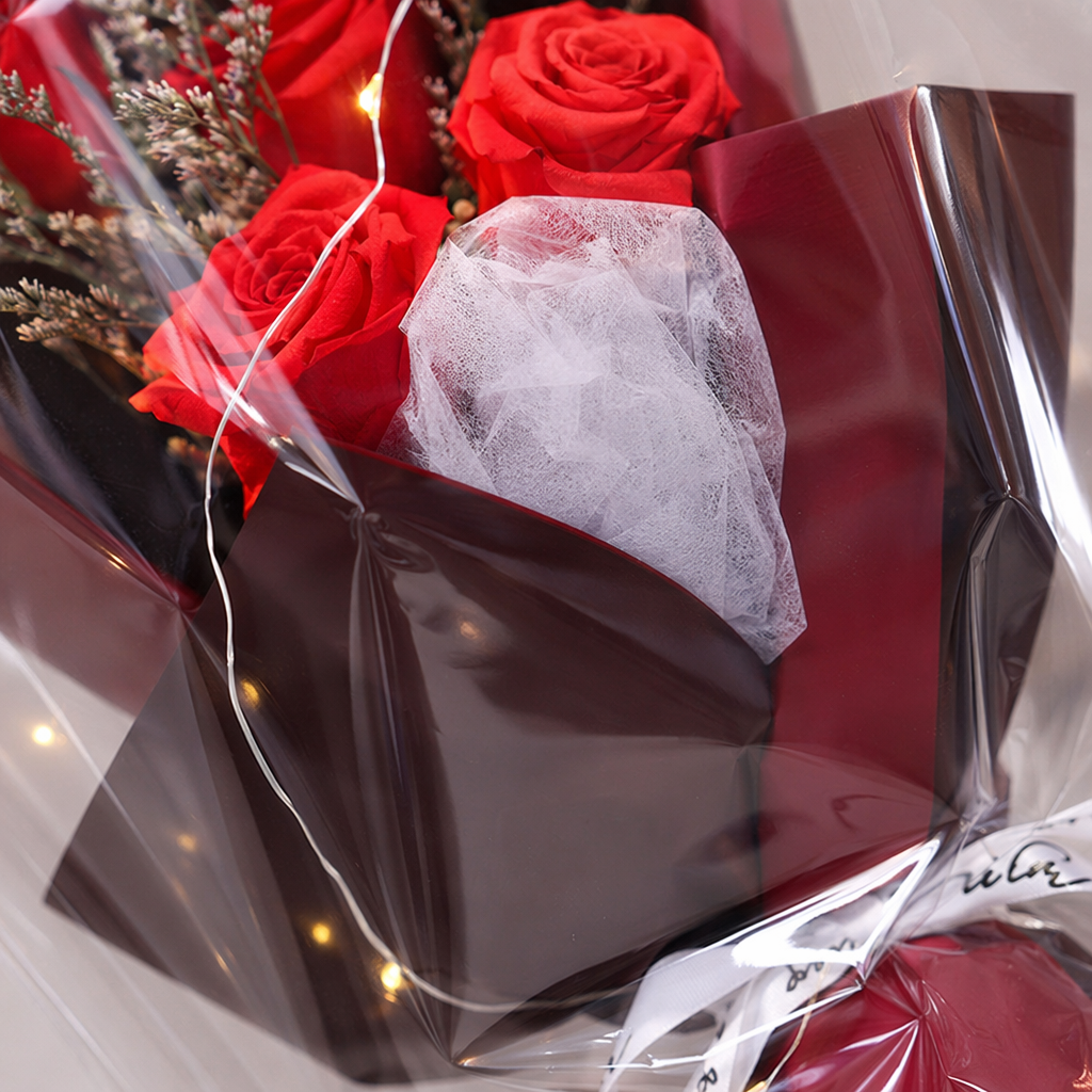 A transparent bagged bouquet of dried red roses with warm light strings, wrapped in red and black paper and white ribbon gift box Application Scenario