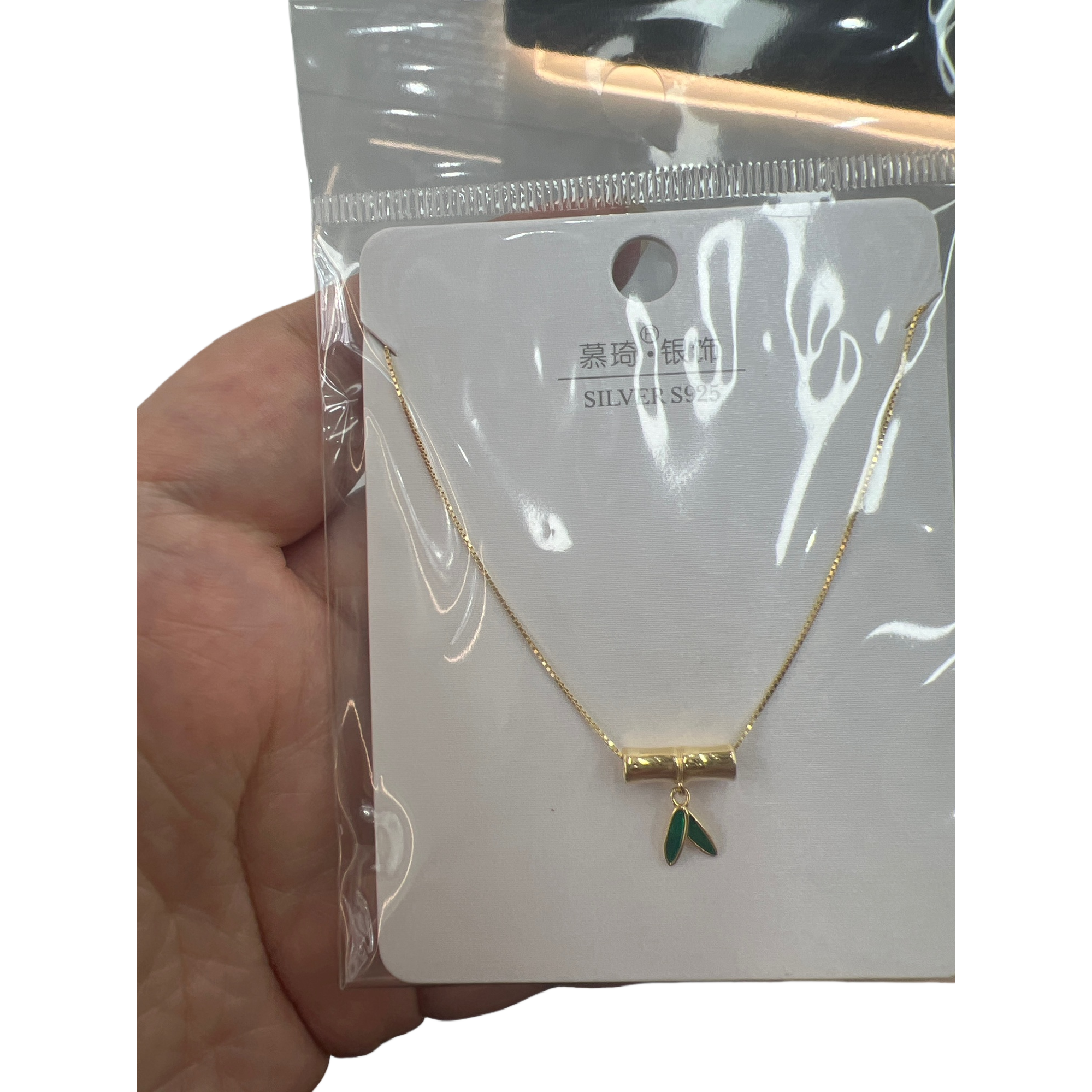 A golden bamboo pendant necklace with green leaves embellished on white cards, in a transparent bag for handheld display Item Picture