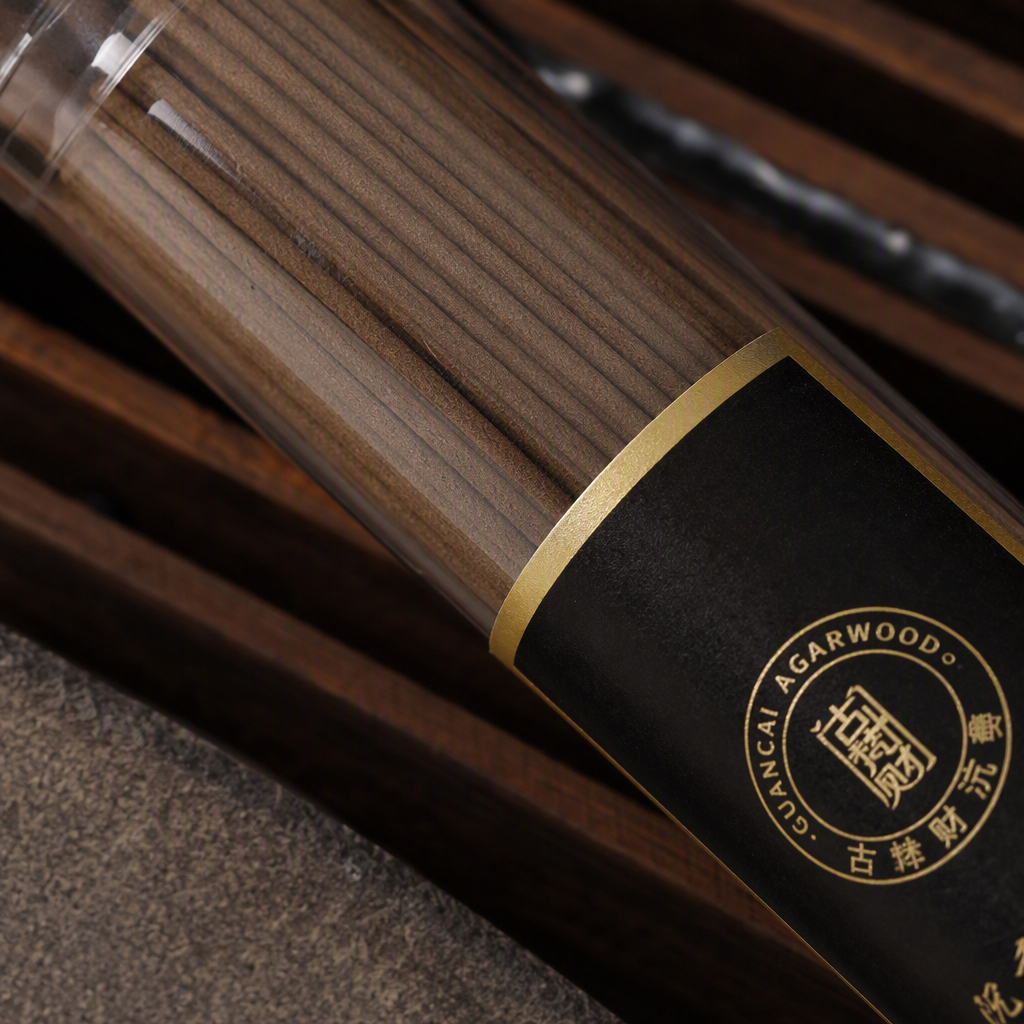 The agarwood incense stick is in a transparent cylinder, with a long brown incense stick. It has a black background and a golden-edge label. The ancient Fan CAI incense burner is used as an indoor fragrance Application Scenario