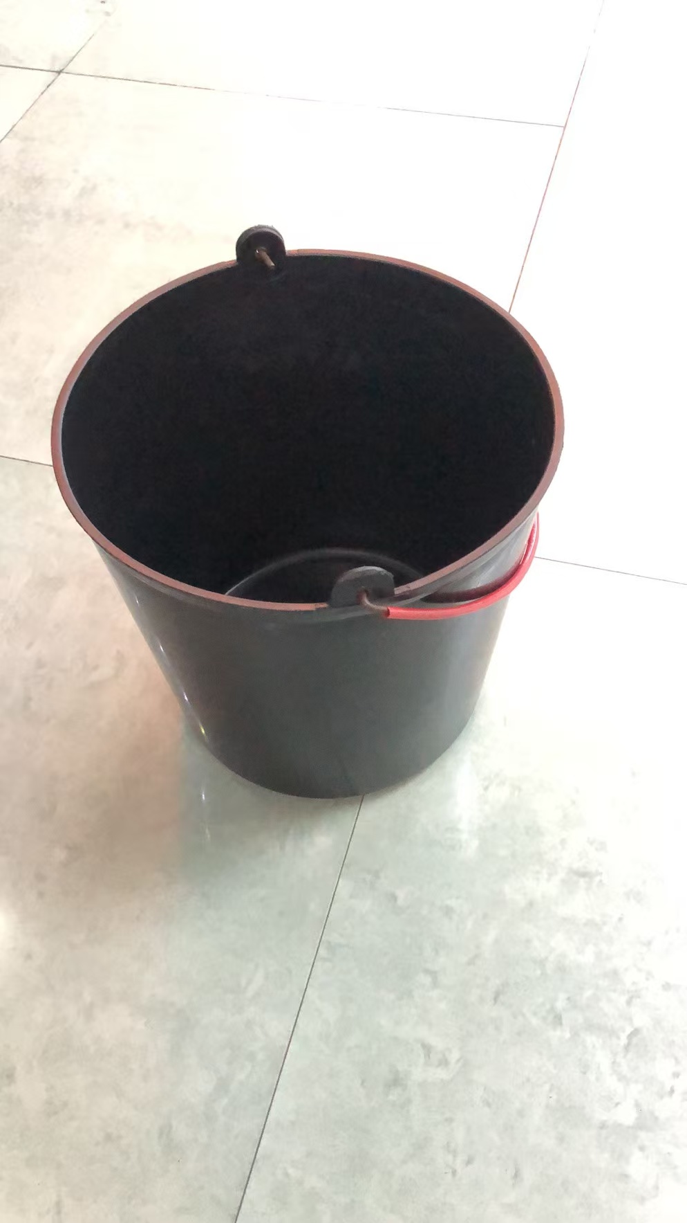 黑色塑料桶，红色提手，置于光滑瓷砖地面，简约实用。水桶塑料水桶