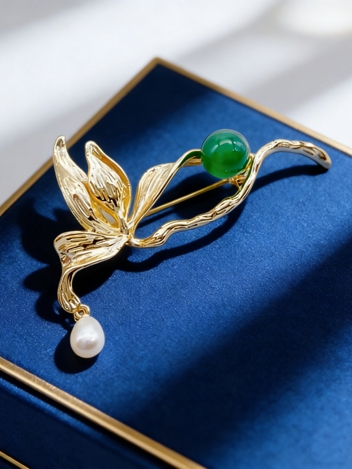 A golden leaf-shaped brooch set with green round beads, white pearls and a fashionable pin in a blue velvet box